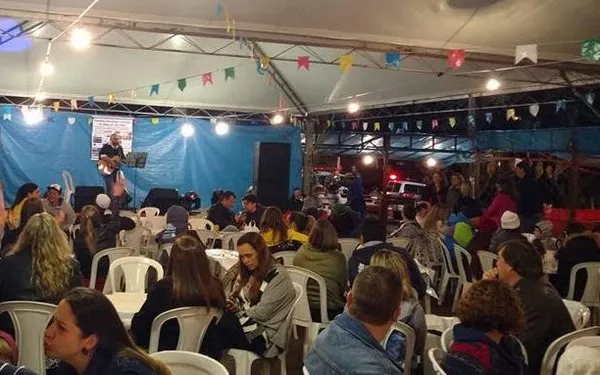 Pessoas sentadas em mesas plásticas sob uma tenda decorada com bandeirinhas coloridas, durante evento noturno com música ao vivo