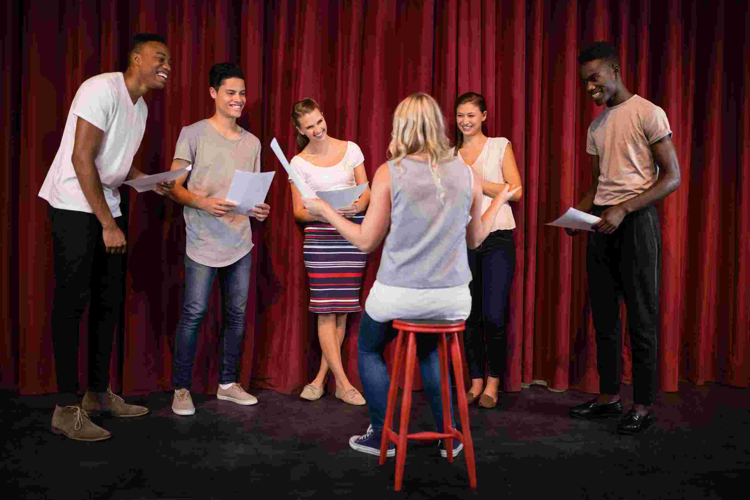 Grupo de atores em pé segurando roteiros e sorrindo durante ensaio de teatro, com uma diretora sentada em um banquinho vermelho de frente para eles, em um palco com cortinas vermelhas ao fundo